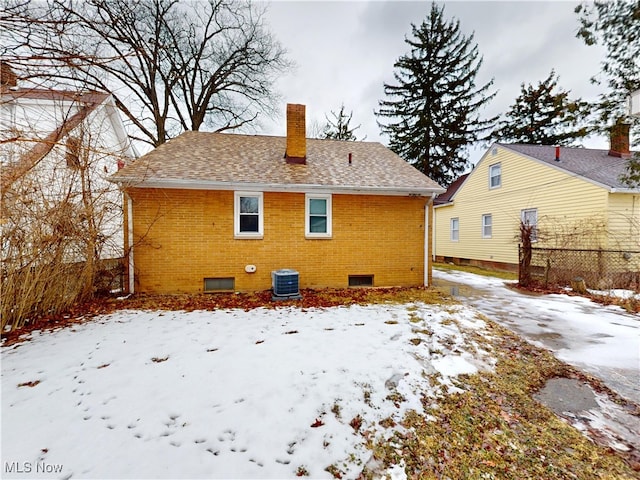 snow covered house featuring brick siding, roof with shingles, a chimney, and central AC unit