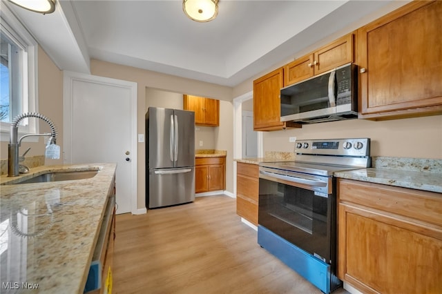 kitchen with brown cabinets, light wood-style flooring, appliances with stainless steel finishes, a sink, and light stone countertops