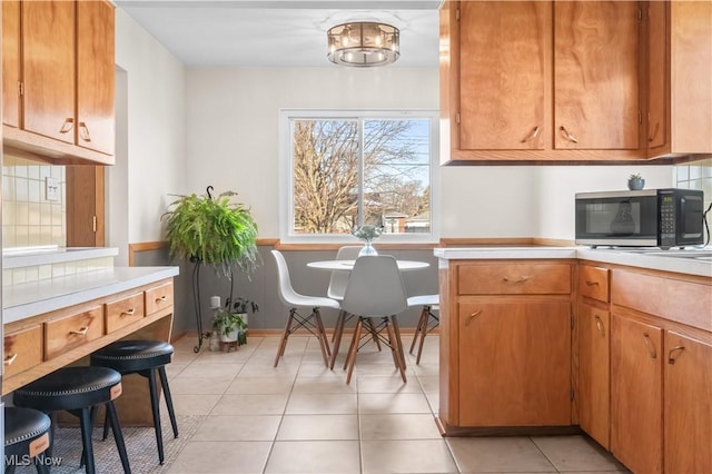 kitchen featuring stainless steel microwave, light countertops, light tile patterned floors, decorative backsplash, and brown cabinets