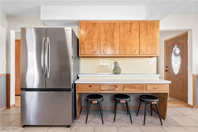 kitchen featuring backsplash, light countertops, light tile patterned floors, freestanding refrigerator, and brown cabinetry