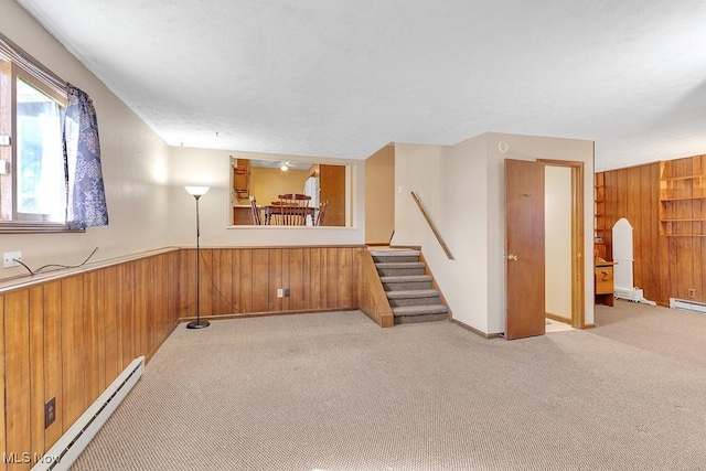 carpeted empty room with wood walls, stairs, a baseboard heating unit, and wainscoting