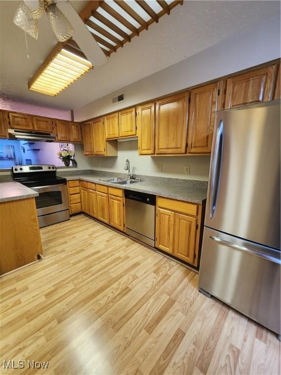 kitchen with light wood finished floors, appliances with stainless steel finishes, brown cabinetry, a sink, and under cabinet range hood