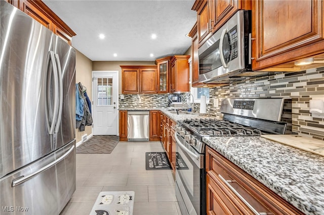 kitchen featuring light stone counters, stainless steel appliances, decorative backsplash, brown cabinetry, and glass insert cabinets