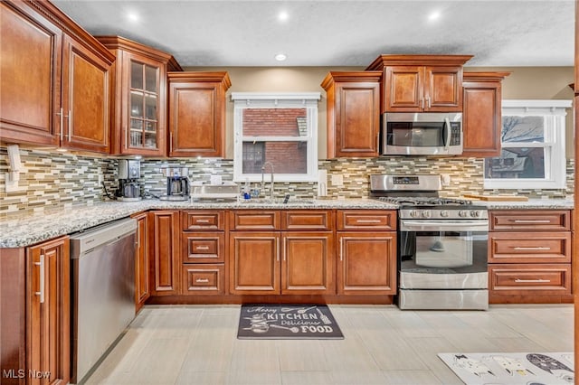 kitchen featuring glass insert cabinets, appliances with stainless steel finishes, backsplash, light stone counters, and a sink