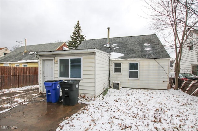 snow covered house with a shingled roof, cooling unit, and fence