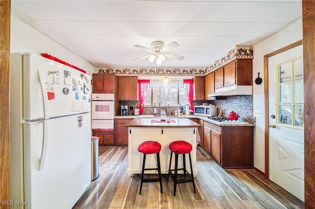 kitchen with under cabinet range hood, white appliances, wood finished floors, backsplash, and a warming drawer