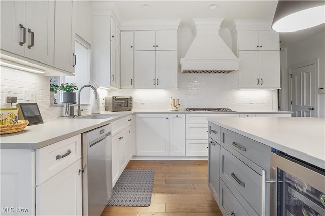 kitchen featuring custom exhaust hood, light countertops, stainless steel dishwasher, light wood-style floors, and beverage cooler