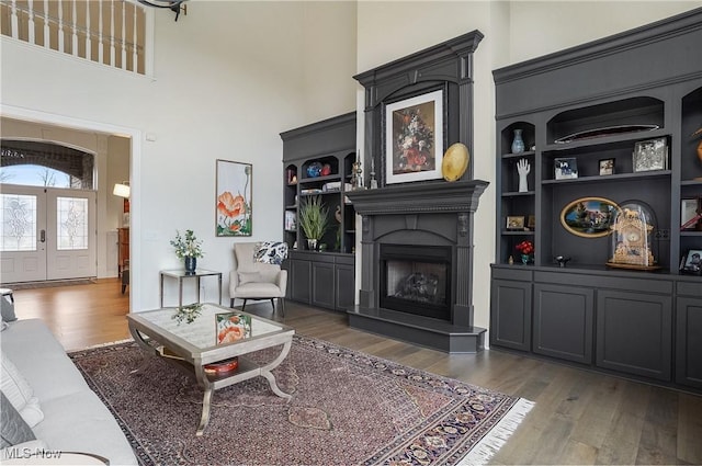 living room featuring built in features, french doors, a fireplace with raised hearth, a high ceiling, and wood finished floors