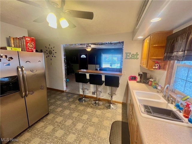 kitchen featuring stainless steel refrigerator with ice dispenser, light countertops, brown cabinetry, a sink, and baseboards