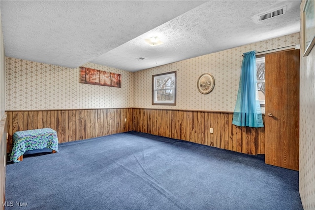 empty room featuring a textured ceiling, a wainscoted wall, visible vents, carpet, and wallpapered walls