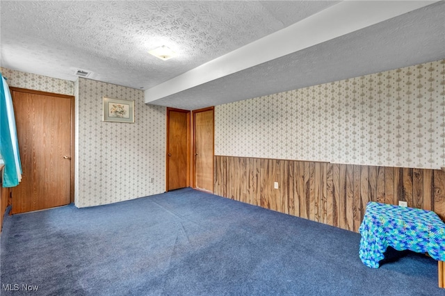 carpeted empty room with a wainscoted wall, visible vents, wood walls, a textured ceiling, and wallpapered walls