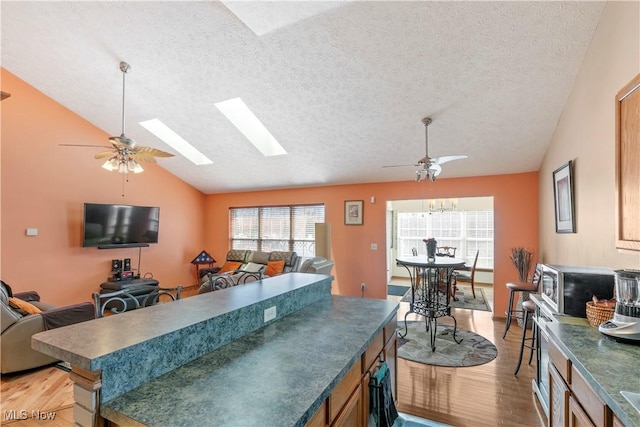 kitchen featuring vaulted ceiling with skylight, light wood-style flooring, a ceiling fan, open floor plan, and dark countertops