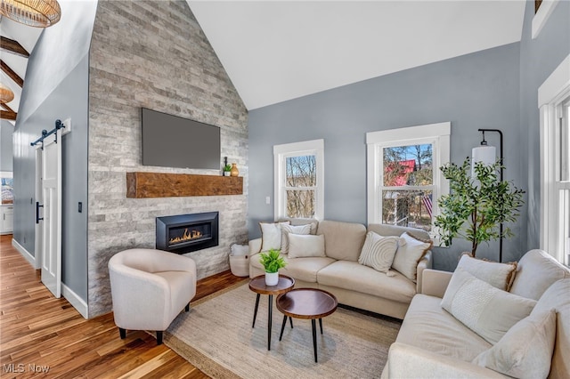 living room featuring a barn door, light wood-style floors, a stone fireplace, high vaulted ceiling, and baseboards