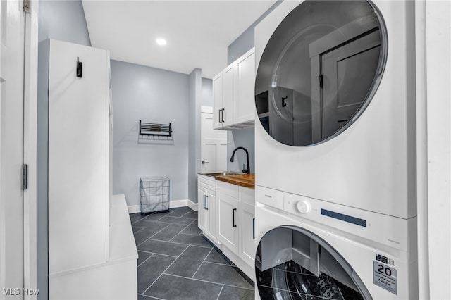 clothes washing area featuring stacked washer and dryer, dark tile patterned flooring, a sink, baseboards, and cabinet space