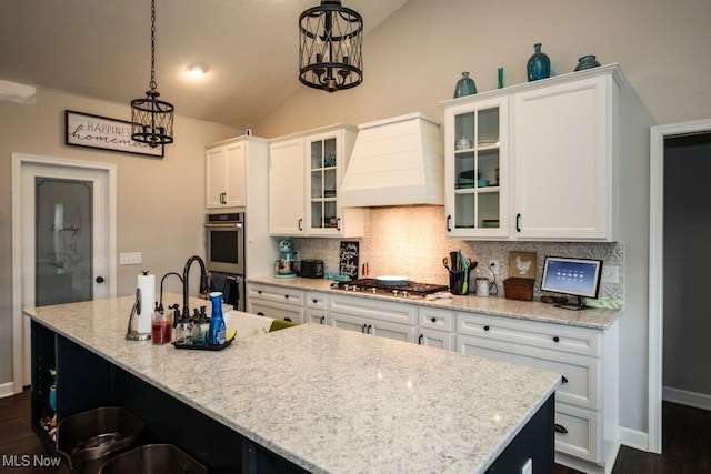 kitchen with an island with sink, custom range hood, vaulted ceiling, decorative backsplash, and appliances with stainless steel finishes