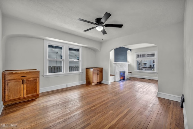 unfurnished living room with ceiling fan, arched walkways, hardwood / wood-style flooring, baseboards, and a glass covered fireplace