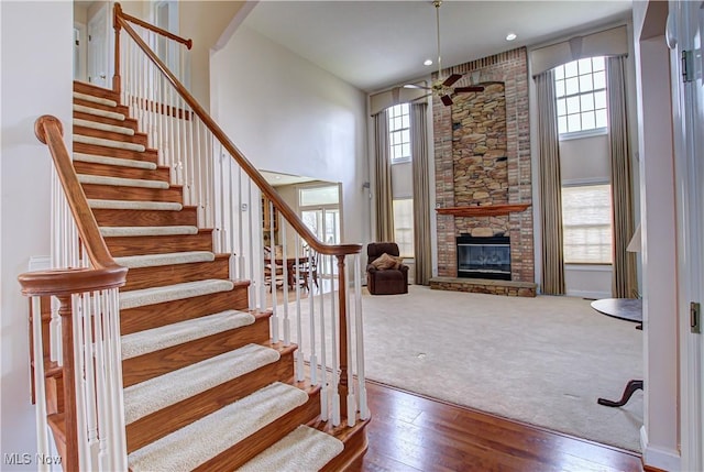 staircase featuring a fireplace, a towering ceiling, a ceiling fan, carpet flooring, and hardwood / wood-style floors