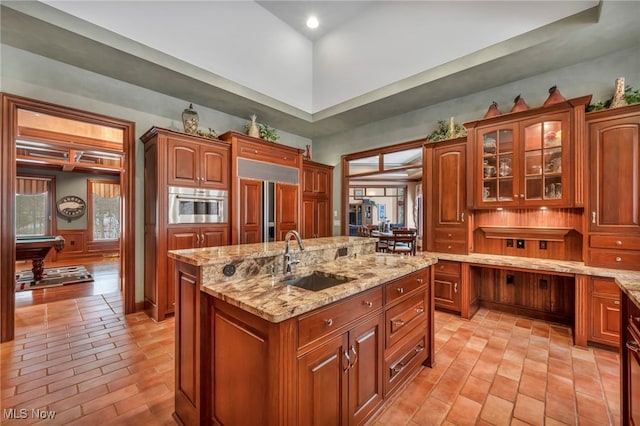 kitchen featuring a sink, oven, glass insert cabinets, light stone counters, and a kitchen island with sink