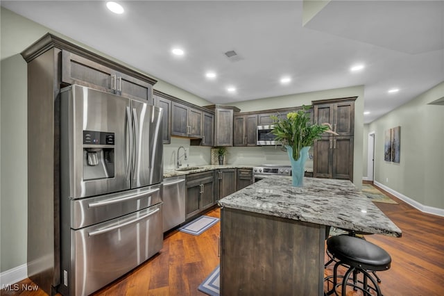 kitchen with light stone countertops, dark wood-style floors, a sink, dark brown cabinetry, and appliances with stainless steel finishes