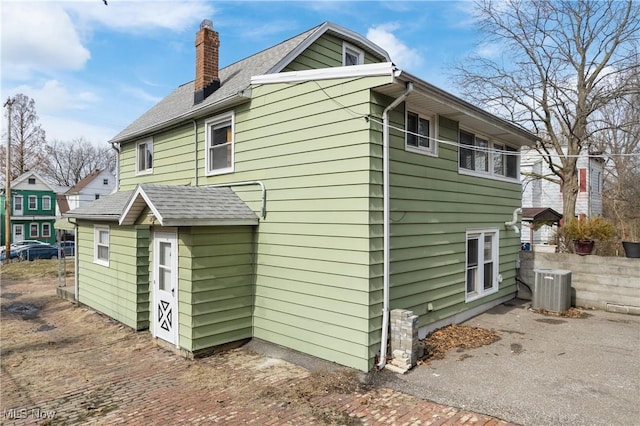 back of house featuring central AC, a shingled roof, and a chimney