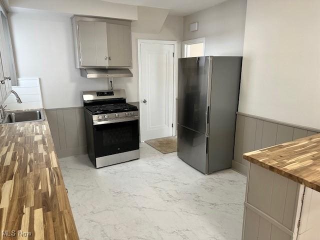 kitchen featuring marble finish floor, appliances with stainless steel finishes, wainscoting, a sink, and wood counters