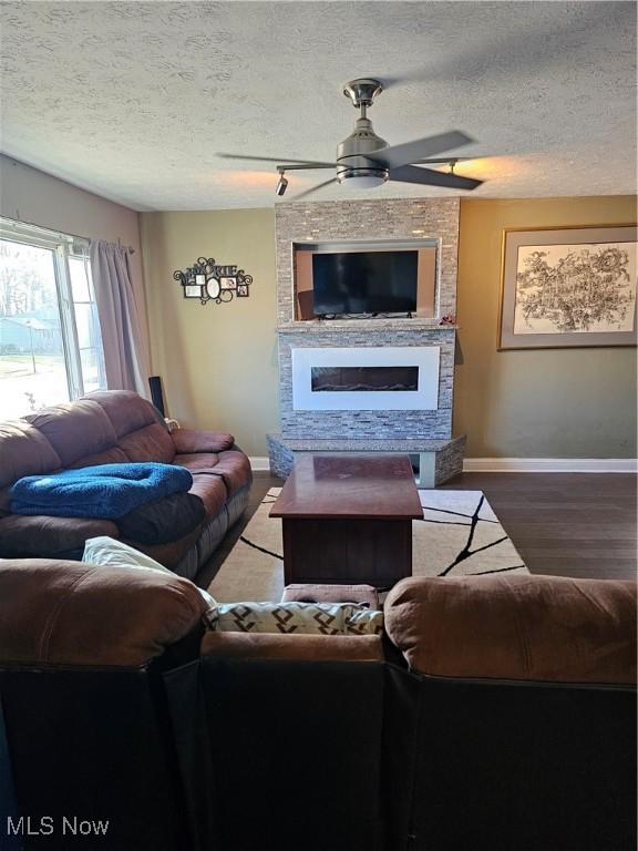 living area featuring a ceiling fan, a textured ceiling, wood finished floors, a large fireplace, and baseboards