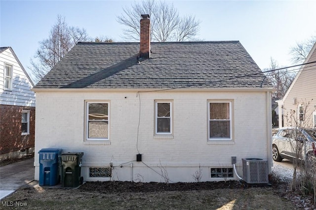 rear view of house featuring a shingled roof, a chimney, central AC unit, and brick siding