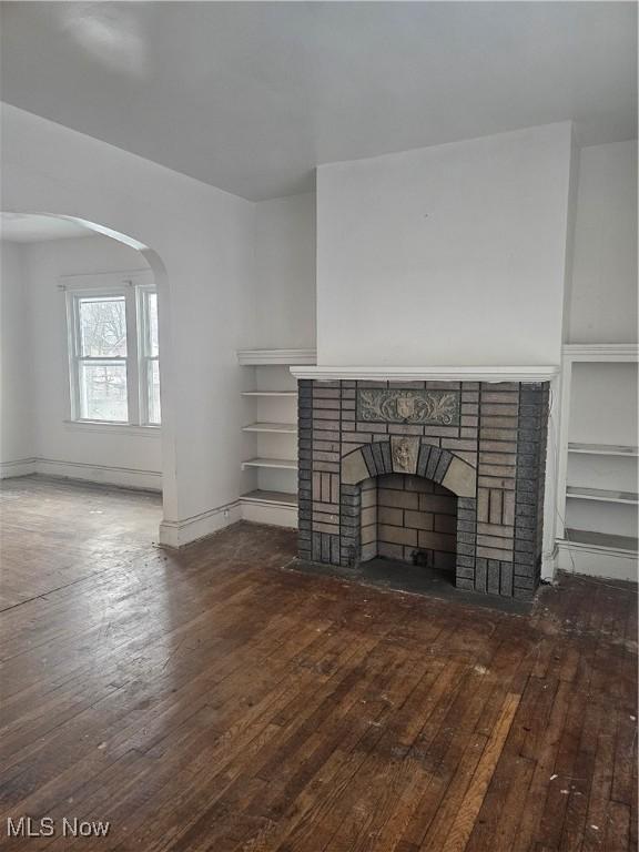 unfurnished living room featuring baseboards, arched walkways, a brick fireplace, and hardwood / wood-style flooring