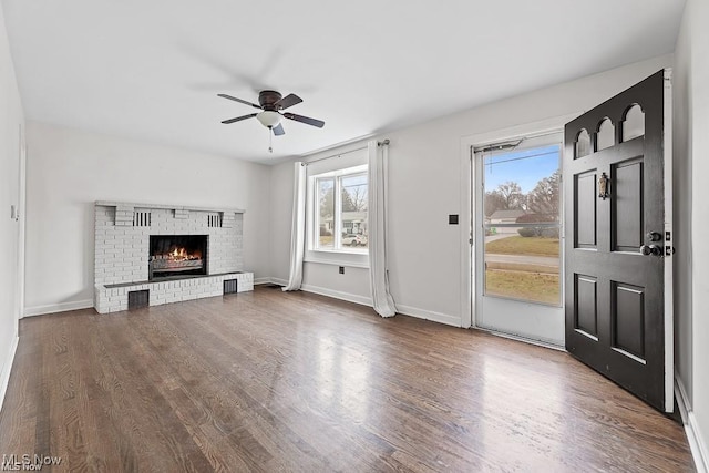 unfurnished living room featuring ceiling fan, dark wood-style flooring, a brick fireplace, and baseboards