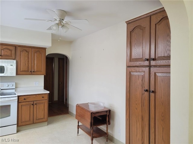 kitchen featuring arched walkways, white appliances, a ceiling fan, light countertops, and brown cabinetry