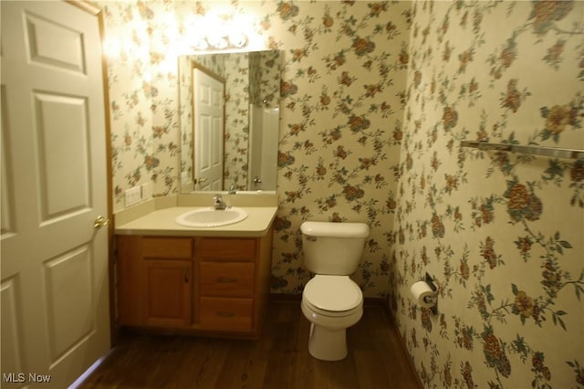 half bathroom with toilet, vanity, wood finished floors, baseboards, and wallpapered walls