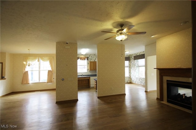 unfurnished living room with dark wood-style floors, ceiling fan with notable chandelier, a multi sided fireplace, and wallpapered walls