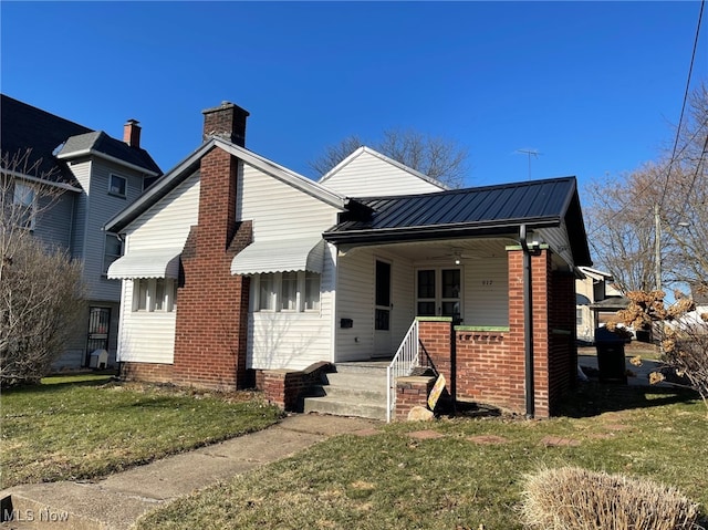 bungalow featuring brick siding, a chimney, covered porch, metal roof, and a front lawn