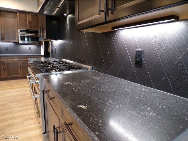 kitchen with ventilation hood, dark stone counters, stainless steel appliances, light wood-style floors, and backsplash