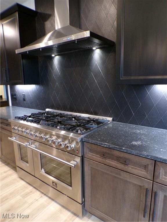 kitchen featuring backsplash, wall chimney range hood, light wood-type flooring, range with two ovens, and dark stone countertops