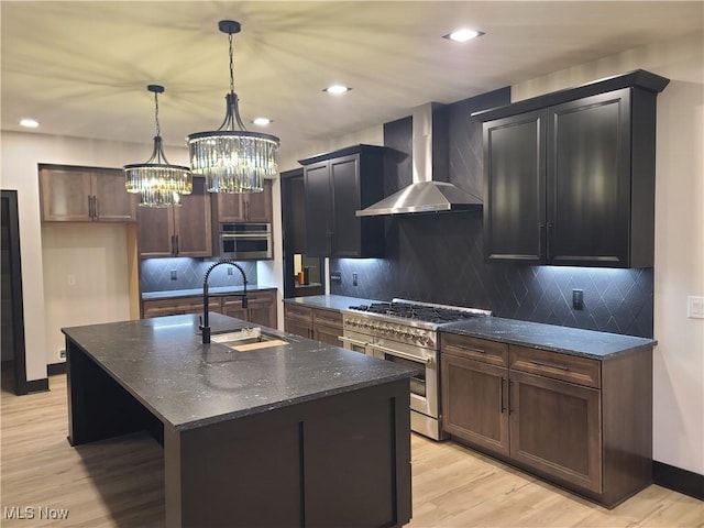 kitchen with a sink, dark stone counters, appliances with stainless steel finishes, wall chimney exhaust hood, and light wood finished floors