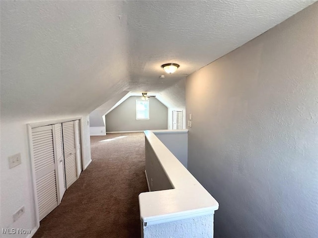 corridor with vaulted ceiling, an upstairs landing, dark colored carpet, and a textured ceiling