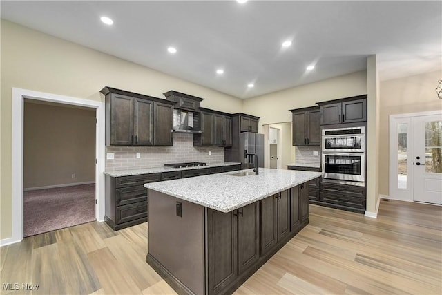 kitchen featuring light wood-style flooring, an island with sink, a sink, appliances with stainless steel finishes, and wall chimney exhaust hood