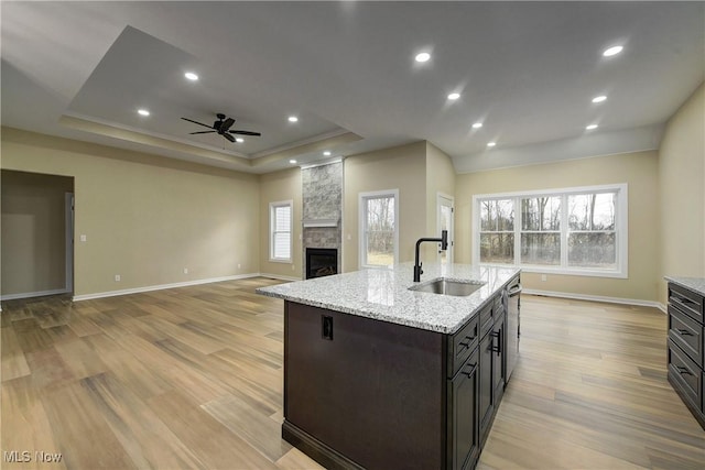 kitchen featuring a large fireplace, an island with sink, a tray ceiling, light wood-style flooring, and a sink