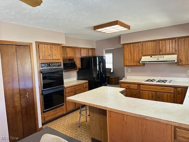 kitchen with under cabinet range hood, brown cabinets, and black appliances