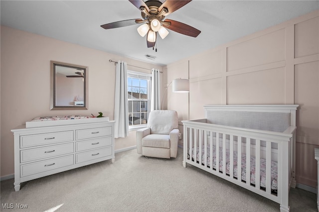 bedroom with visible vents, a crib, ceiling fan, light colored carpet, and a decorative wall