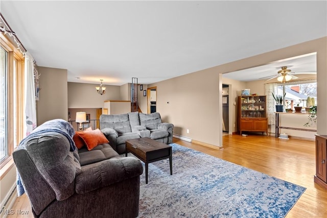 living room with light wood finished floors, ceiling fan with notable chandelier, a baseboard heating unit, and baseboards