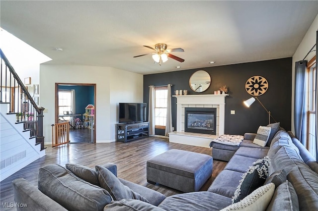 living room featuring dark wood-type flooring, a tiled fireplace, a ceiling fan, stairway, and baseboards