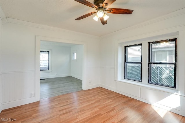 empty room with crown molding, a ceiling fan, light wood-type flooring, and wainscoting