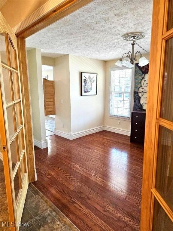 unfurnished dining area with a notable chandelier, a textured ceiling, baseboards, and dark wood-style flooring