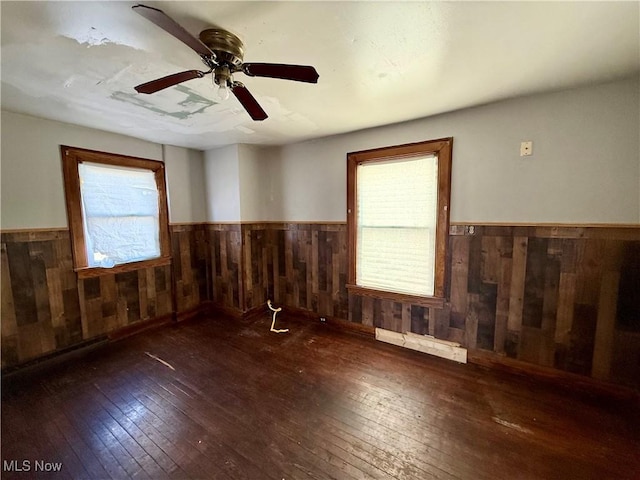 empty room with ceiling fan, wooden walls, wood-type flooring, and wainscoting