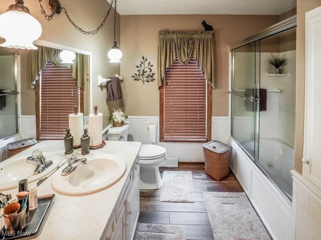 bathroom with vanity, wood finished floors, bath / shower combo with glass door, wainscoting, and toilet