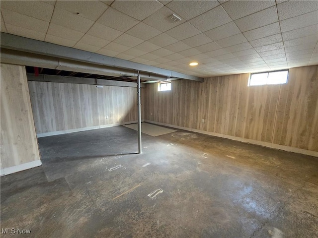 basement with wooden walls, a paneled ceiling, baseboards, and a healthy amount of sunlight