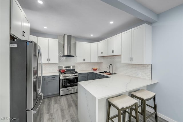 kitchen with a peninsula, a sink, stainless steel appliances, light wood-style floors, and wall chimney exhaust hood