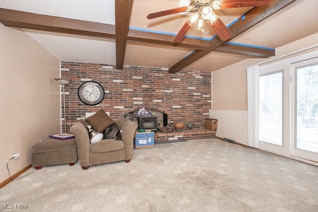 unfurnished living room featuring a wainscoted wall, a wood stove, ceiling fan, beamed ceiling, and carpet flooring